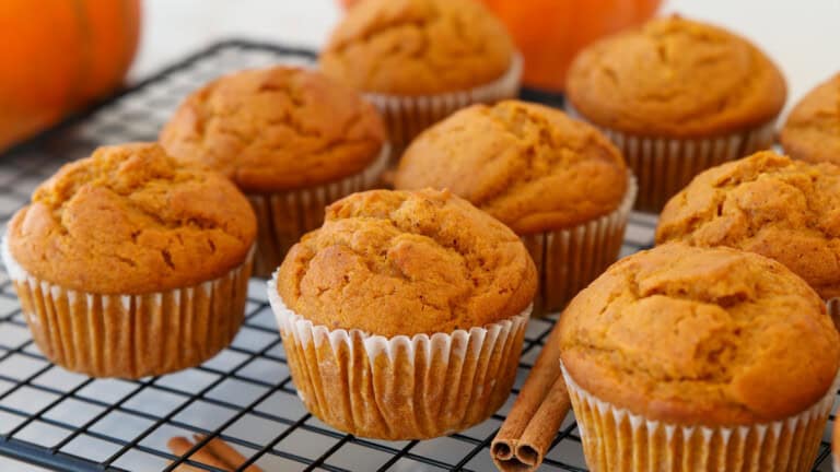 Pumpkin muffins in white paper liners cooling on a black wire rack, with a cinnamon stick and pumpkins blurred in the background.