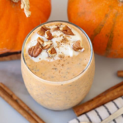 A glass of pumpkin overnight oats topped with whipped cream, chopped pecans, and a sprinkle of spices, surrounded by whole pumpkins and cinnamon sticks on a white surface.
