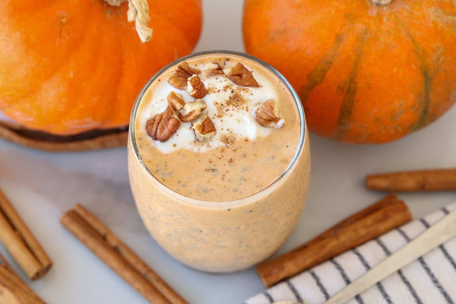 A glass of pumpkin overnight oats topped with whipped cream, chopped pecans, and a sprinkle of spices, surrounded by whole pumpkins and cinnamon sticks on a white surface.