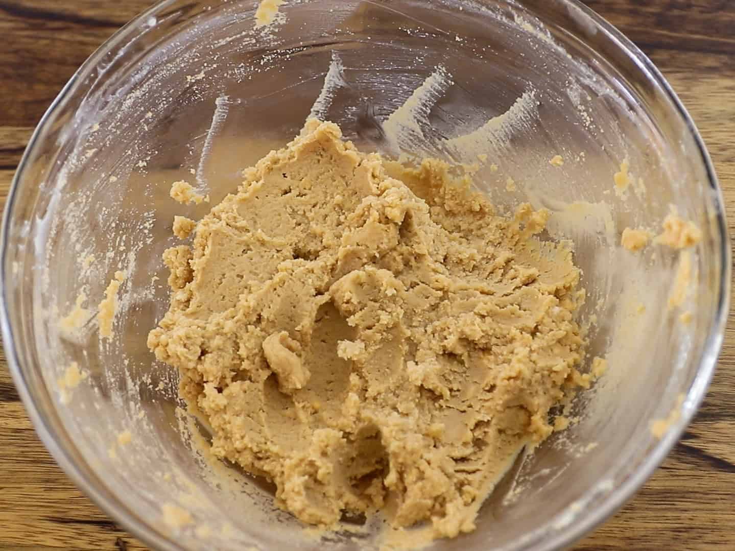 A glass bowl containing creamy, partially mixed cookie dough sits on a wooden surface. The dough is a light beige color and is pressed against the sides of the bowl.