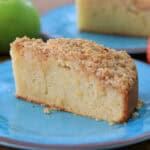 A slice of crumb-topped apple cake sits on a blue plate, with whole green and red apples and more cake visible in the background.