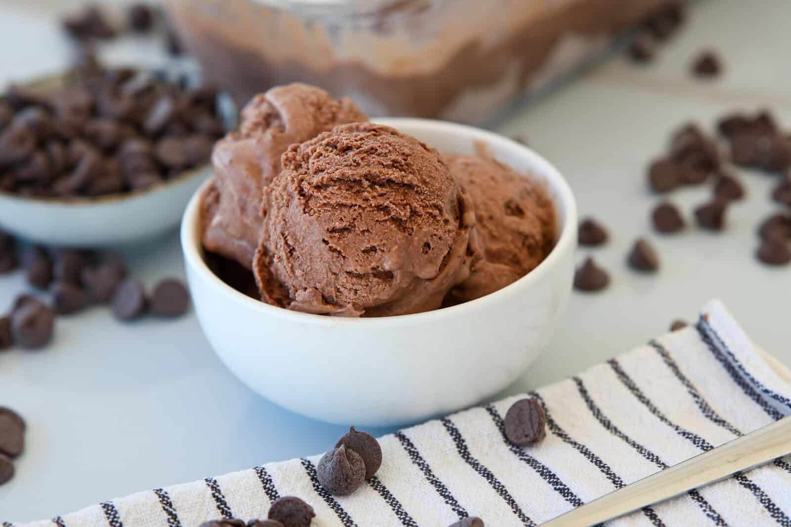 A white bowl filled with scoops of chocolate ice cream sits on a striped cloth, surrounded by scattered chocolate chips and a spoon. A bowl and tray of more chocolate chips and ice cream are visible in the background.