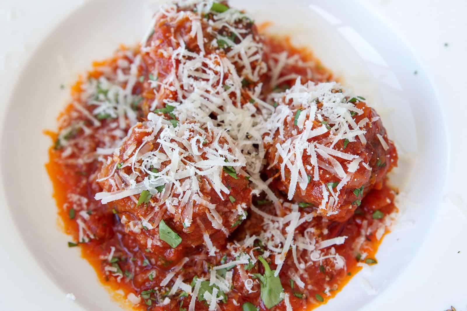 Three large meatballs covered in tomato sauce, garnished with chopped herbs and generously topped with grated parmesan cheese, served on a white plate.