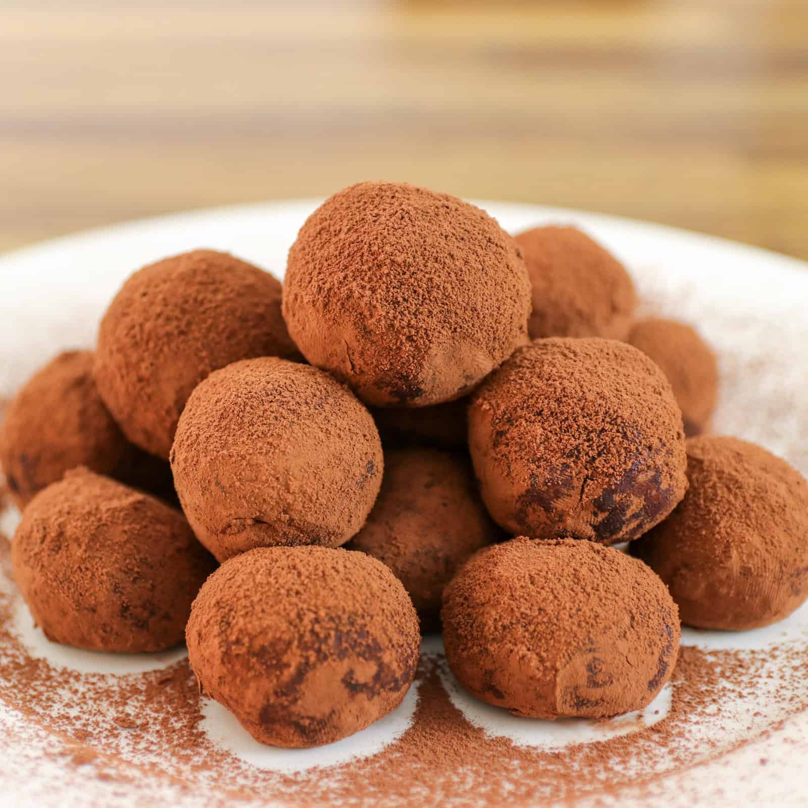 A pile of chocolate truffles dusted with cocoa powder is arranged on a white plate, with a blurred wooden surface in the background.