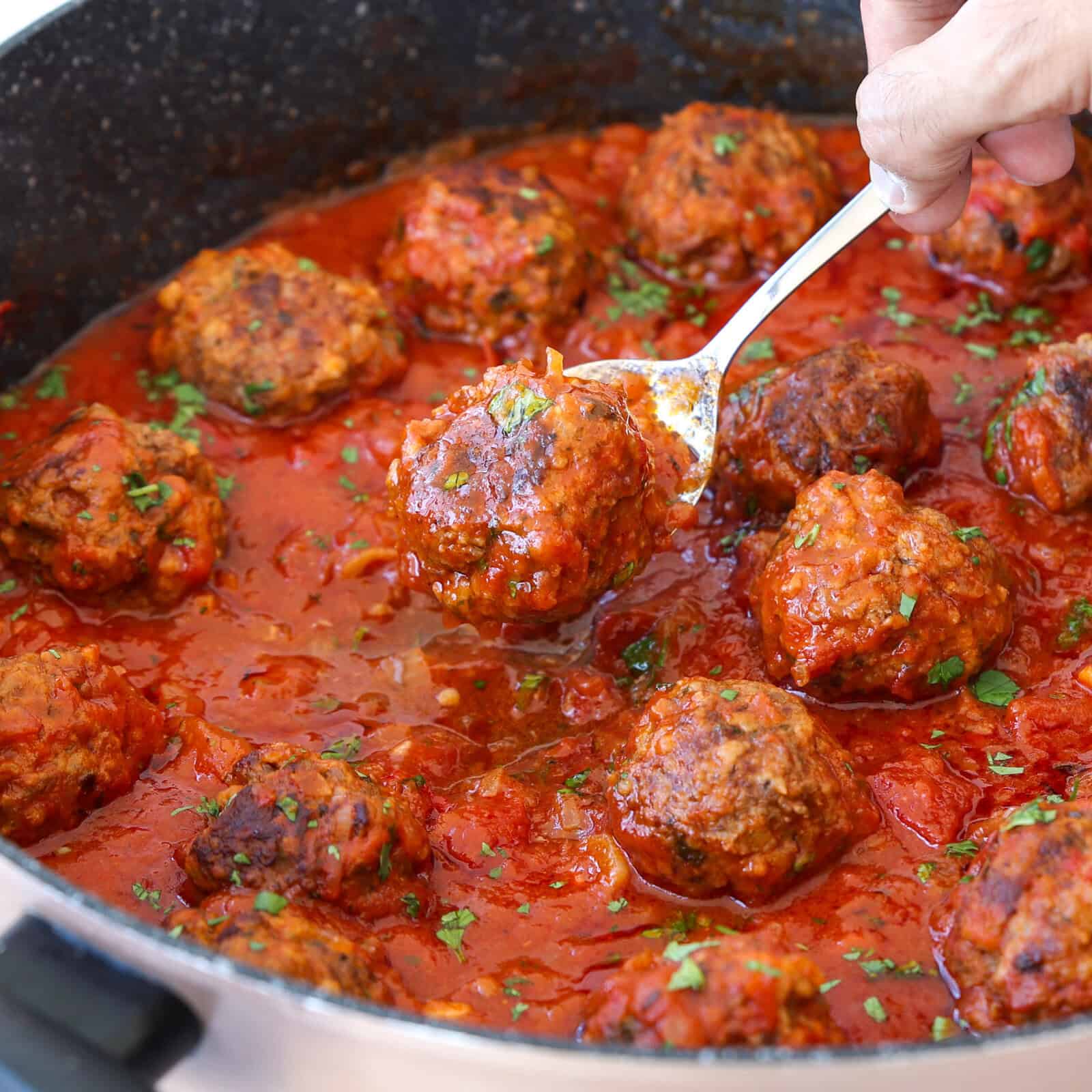 A hand holds a spoon lifting a meatball from a pan filled with meatballs in red tomato sauce, garnished with chopped herbs.