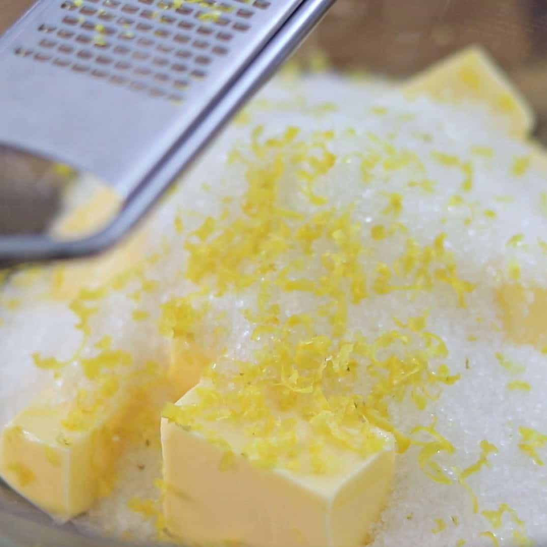 A metal grater rests above a bowl containing sugar, cubed butter, and freshly grated lemon zest.