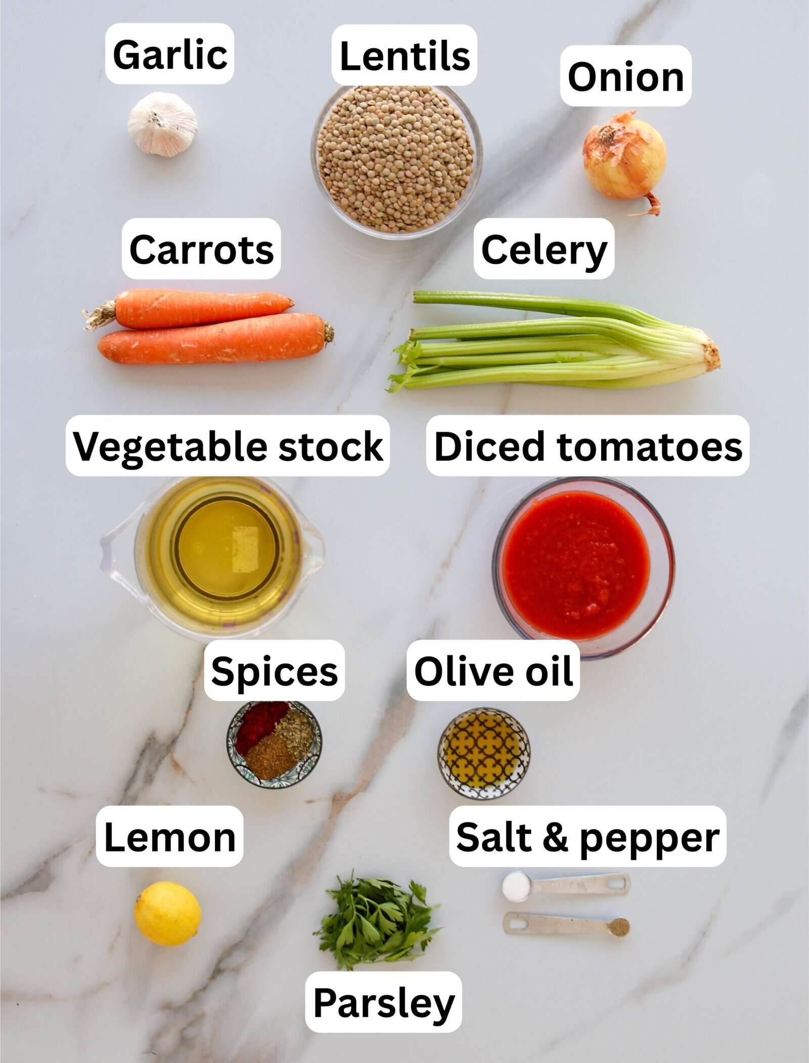 Top-down view of labeled ingredients for a lentil soup: garlic, lentils, onion, carrots, celery, vegetable stock, diced tomatoes, spices, olive oil, lemon, salt & pepper, and parsley, arranged on a white surface.