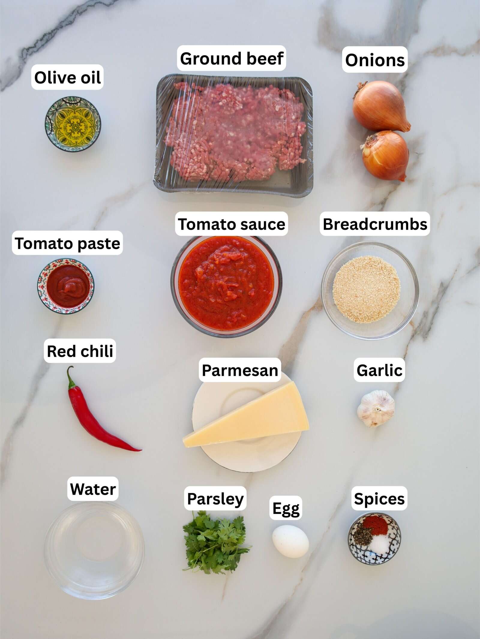 A flat lay of labeled ingredients on a marble surface, including ground beef, onions, olive oil, tomato paste, tomato sauce, breadcrumbs, red chili, parmesan, garlic, water, parsley, egg, and spices.