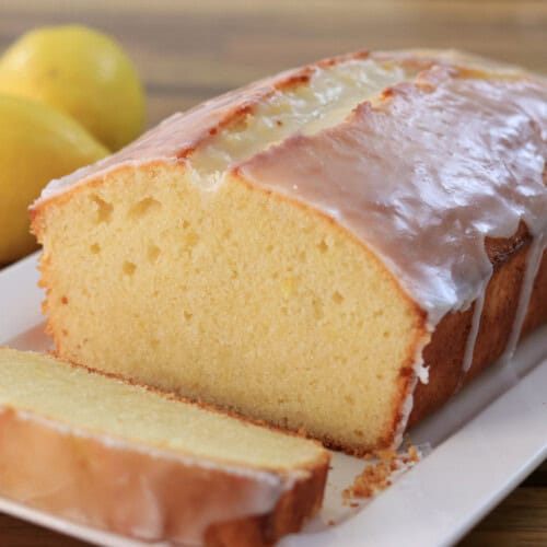 A glazed lemon loaf cake with one slice cut, displayed on a white rectangular plate. Two whole lemons are placed in the background on a wooden surface.