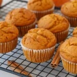 Freshly baked pumpkin muffins in white paper liners cooling on a black wire rack, with cinnamon sticks placed nearby. The muffins are golden brown and have a slightly cracked top.