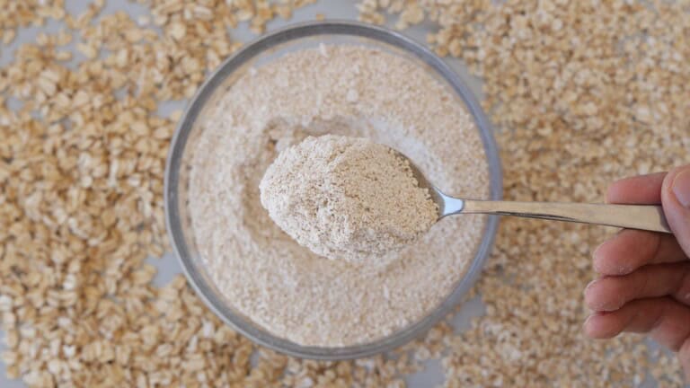 A hand holds a spoonful of fine oat flour above a glass bowl filled with more oat flour, surrounded by scattered rolled oats on a light surface.
