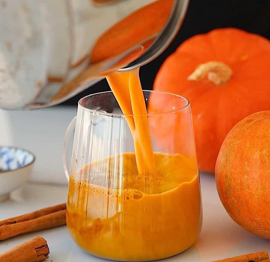 A glass mug is being filled with orange pumpkin spice latte, with whole pumpkins and cinnamon sticks nearby on a white surface.