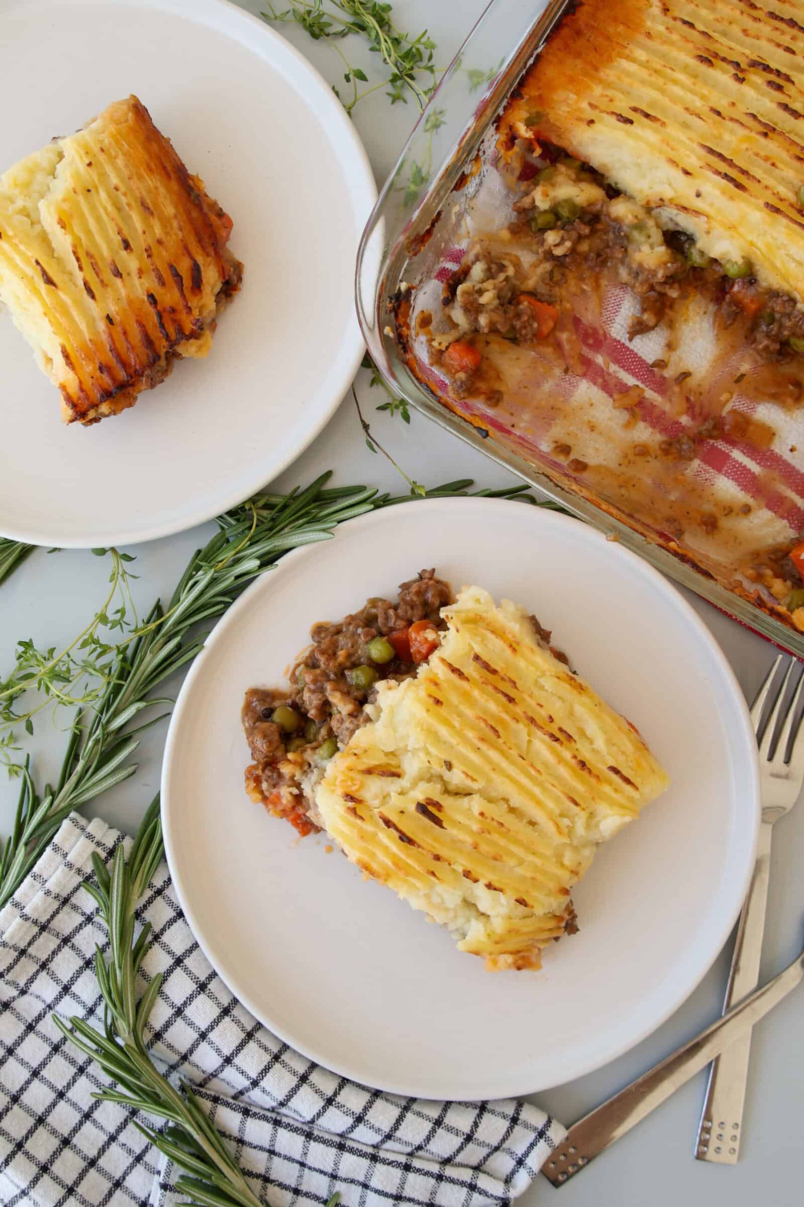 A casserole dish of shepherd’s pie with golden mashed potatoes sits beside two plates, each with a serving. Fresh rosemary and thyme, a fork, knife, and a checkered napkin complete the scene.