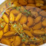 A saucepan filled with peeled garlic cloves submerged in olive oil, garnished with sprigs of fresh thyme and red pepper flakes, sits on a wooden surface.