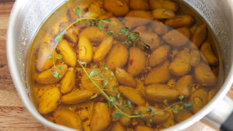 Easy Garlic Confit Recipe | Oven Roasted & Stovetop Methods A saucepan filled with peeled garlic cloves submerged in olive oil, garnished with sprigs of fresh thyme and red pepper flakes, sits on a wooden surface.