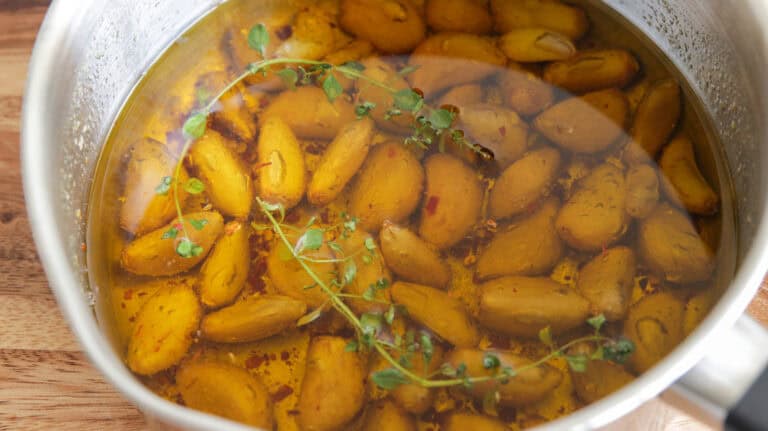 A saucepan filled with peeled garlic cloves submerged in olive oil, garnished with sprigs of fresh thyme and red pepper flakes, sits on a wooden surface.
