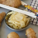 A bowl of thinly sliced pickled ginger surrounded by fresh ginger roots, with a pair of decorative white chopsticks resting on the bowl and a checkered cloth nearby.