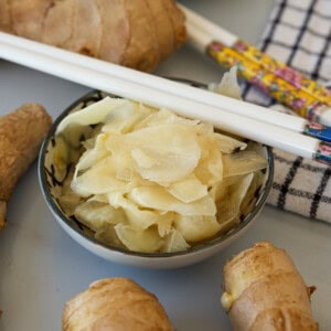 A bowl of thinly sliced pickled ginger surrounded by fresh ginger roots, with a pair of decorative white chopsticks resting on the bowl and a checkered cloth nearby.