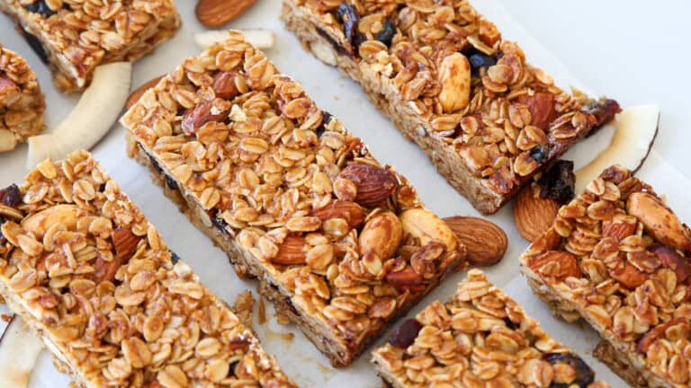 Granola bars with visible oats, almonds, and dried fruit are arranged on parchment paper, surrounded by scattered whole almonds and coconut flakes. The bars look chewy and homemade.