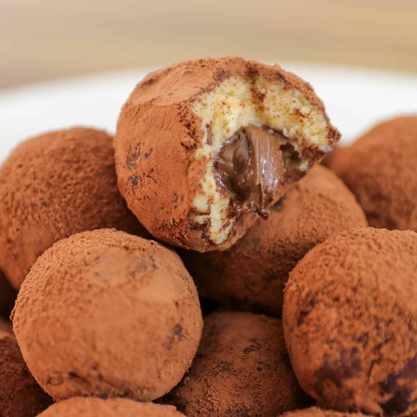 A close-up of several chocolate truffles coated in cocoa powder, with one truffle at the top bitten open to reveal a creamy chocolate filling inside.