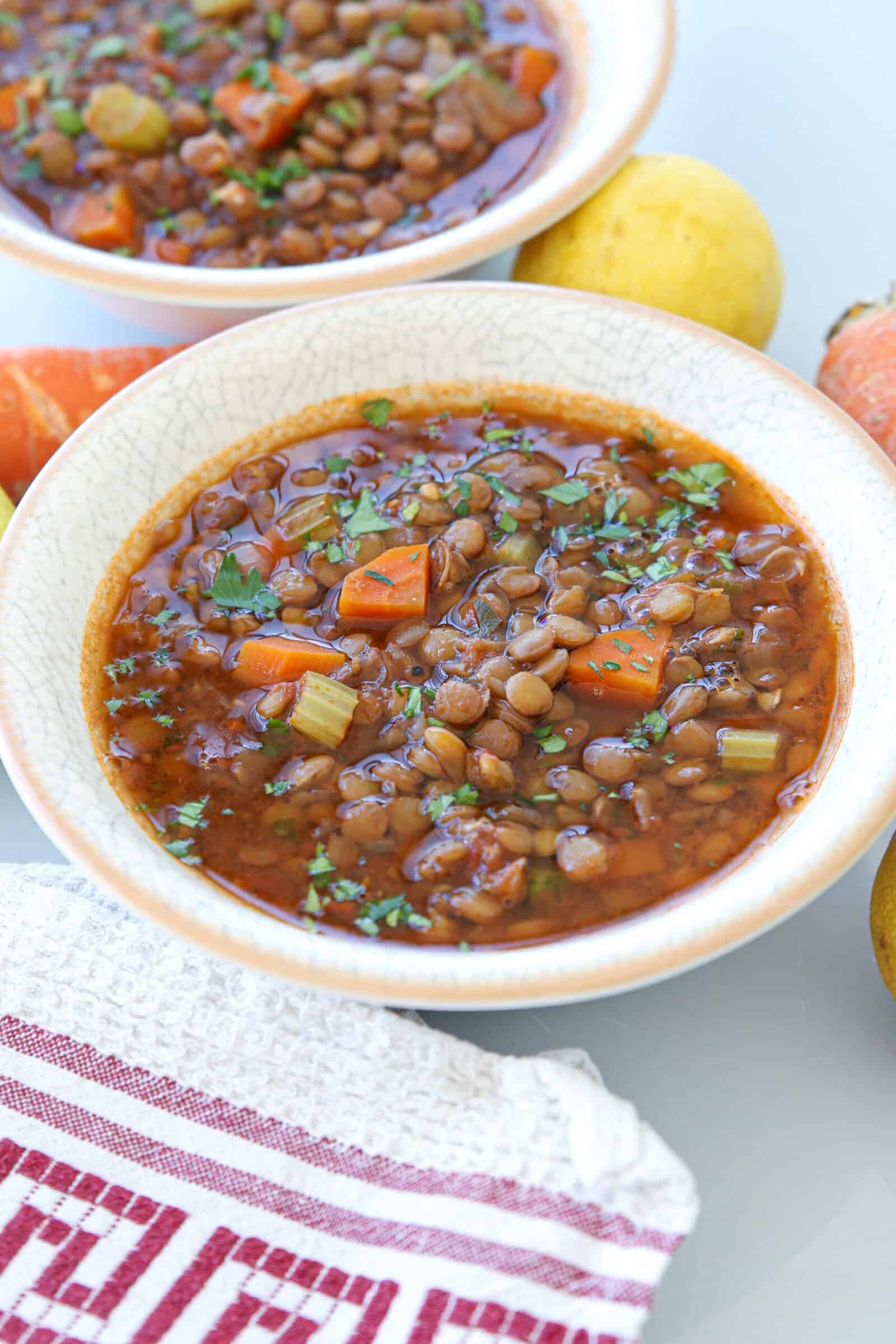 A bowl of lentil soup with chopped carrots, celery, and herbs sits on a table next to a red-striped towel, with another bowl and some whole lemons in the background.