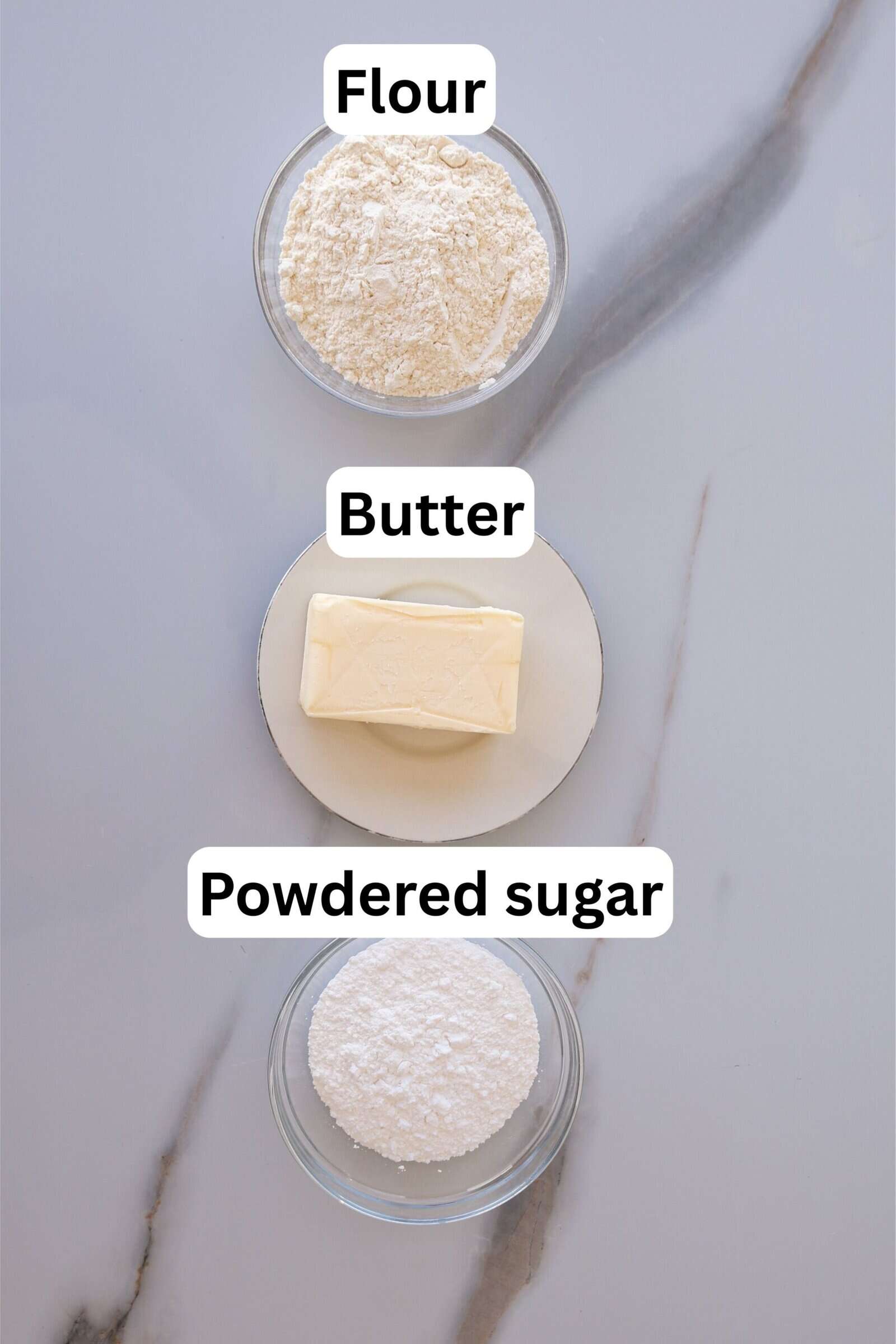 Three labeled bowls on a marble surface: the top bowl contains flour, the middle plate has a stick of butter, and the bottom bowl holds powdered sugar.