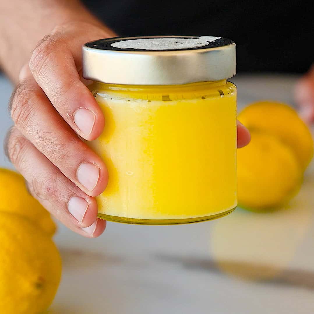A hand holding a glass jar filled with bright yellow lemon curd, with whole lemons visible on a white surface in the background.