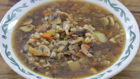 A bowl of mushroom barley soup with sliced mushrooms, barley, carrots, and potatoes in a clear broth, served in a white bowl with a floral pattern on the rim.