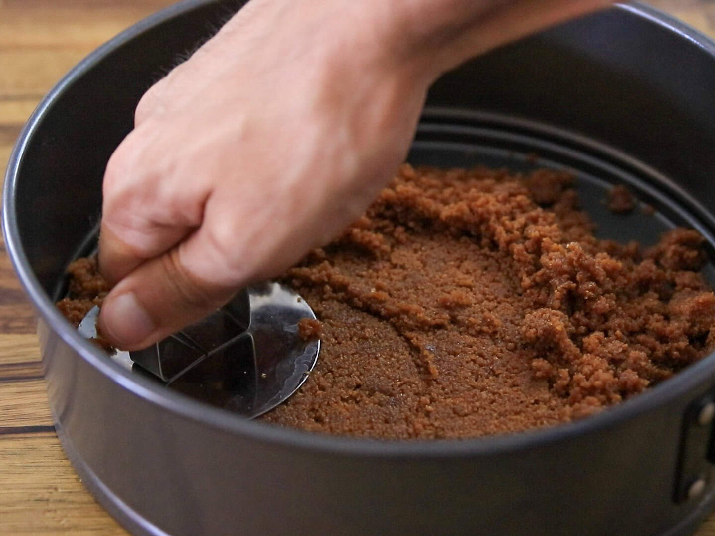 A hand presses a crumbly brown mixture into the bottom of a round springform pan using a metal tool, preparing a crust on a wooden surface.