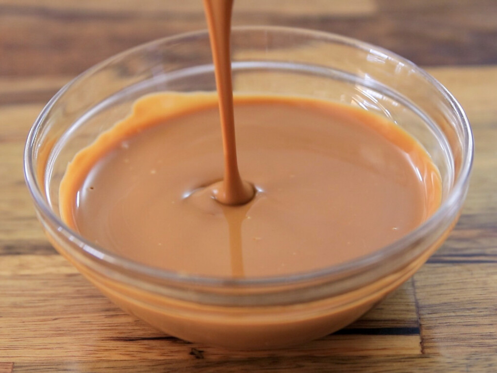 A stream of thick cookie butter is being poured into a clear glass bowl filled with caramel, set on a wooden surface.