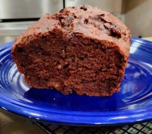 A thick slice of chocolate banana bread rests on a blue plate, showing its rich, moist, dark brown interior with chunks of chocolate throughout.