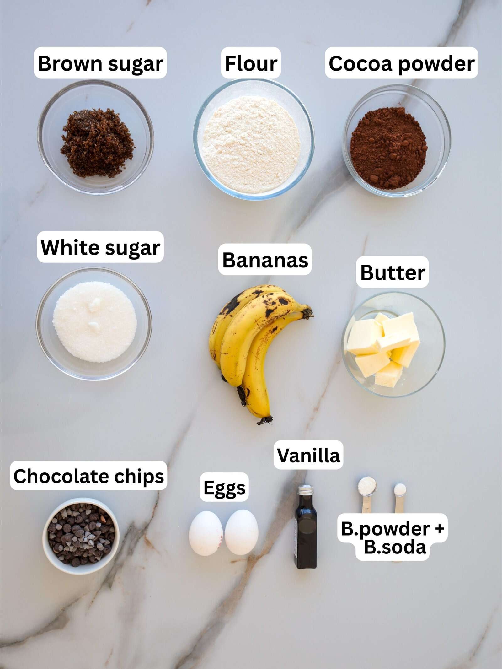 Overhead view of baking ingredients on a white surface: brown sugar, flour, cocoa powder, white sugar, bananas, butter, chocolate chips, eggs, vanilla, baking powder, and baking soda, each labeled.