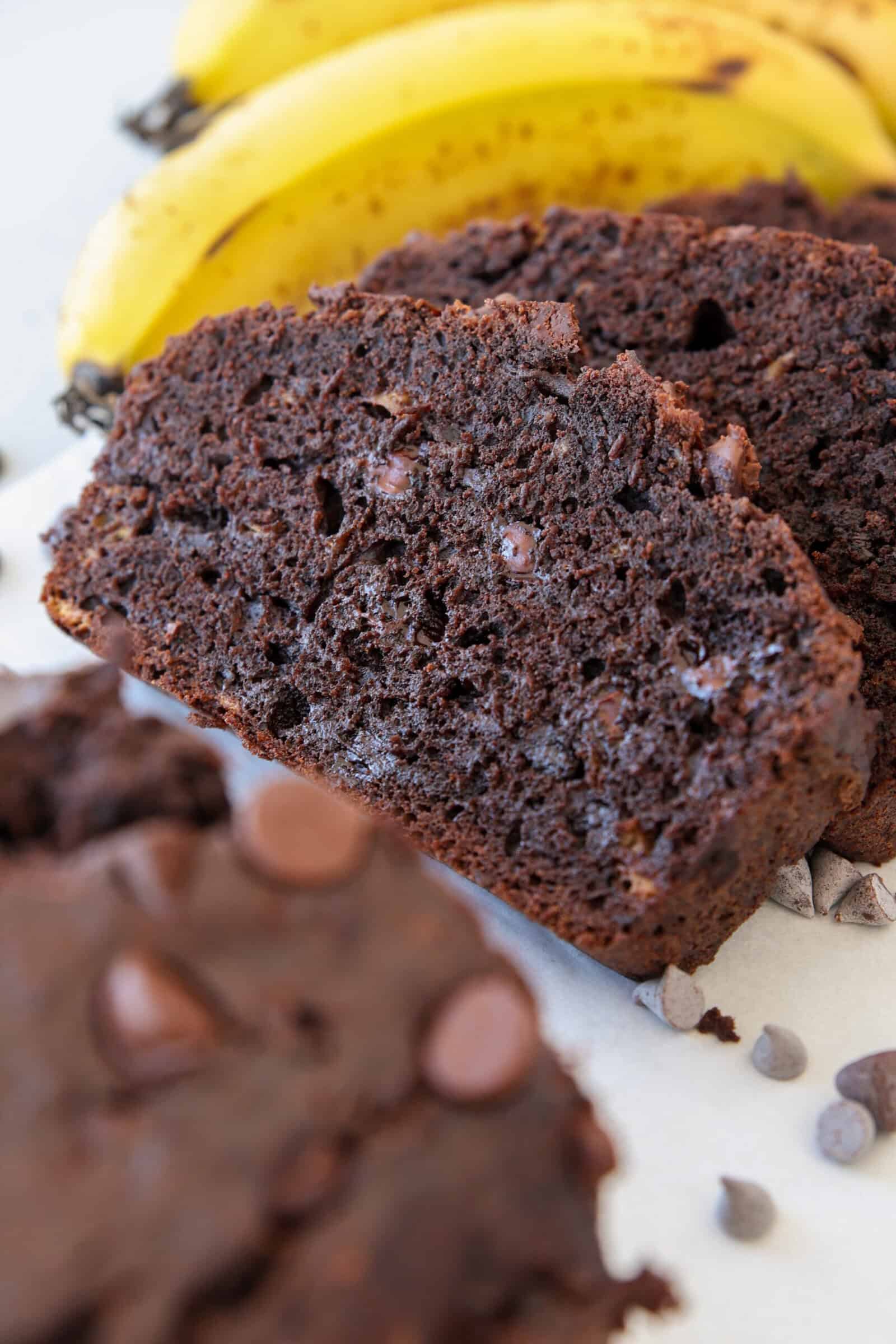 Close-up of two slices of moist chocolate banana bread with chocolate chips, with ripe bananas in the background and chocolate chips scattered nearby.