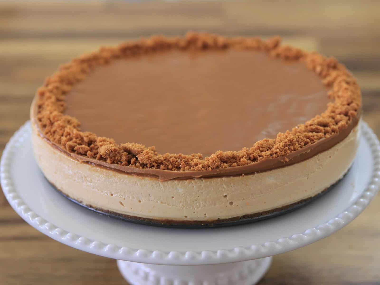 A creamy lotus biscoff cheesecake topped with a smooth caramel glaze and a crumbly cookie crust around the edge, displayed on a white decorative cake stand.