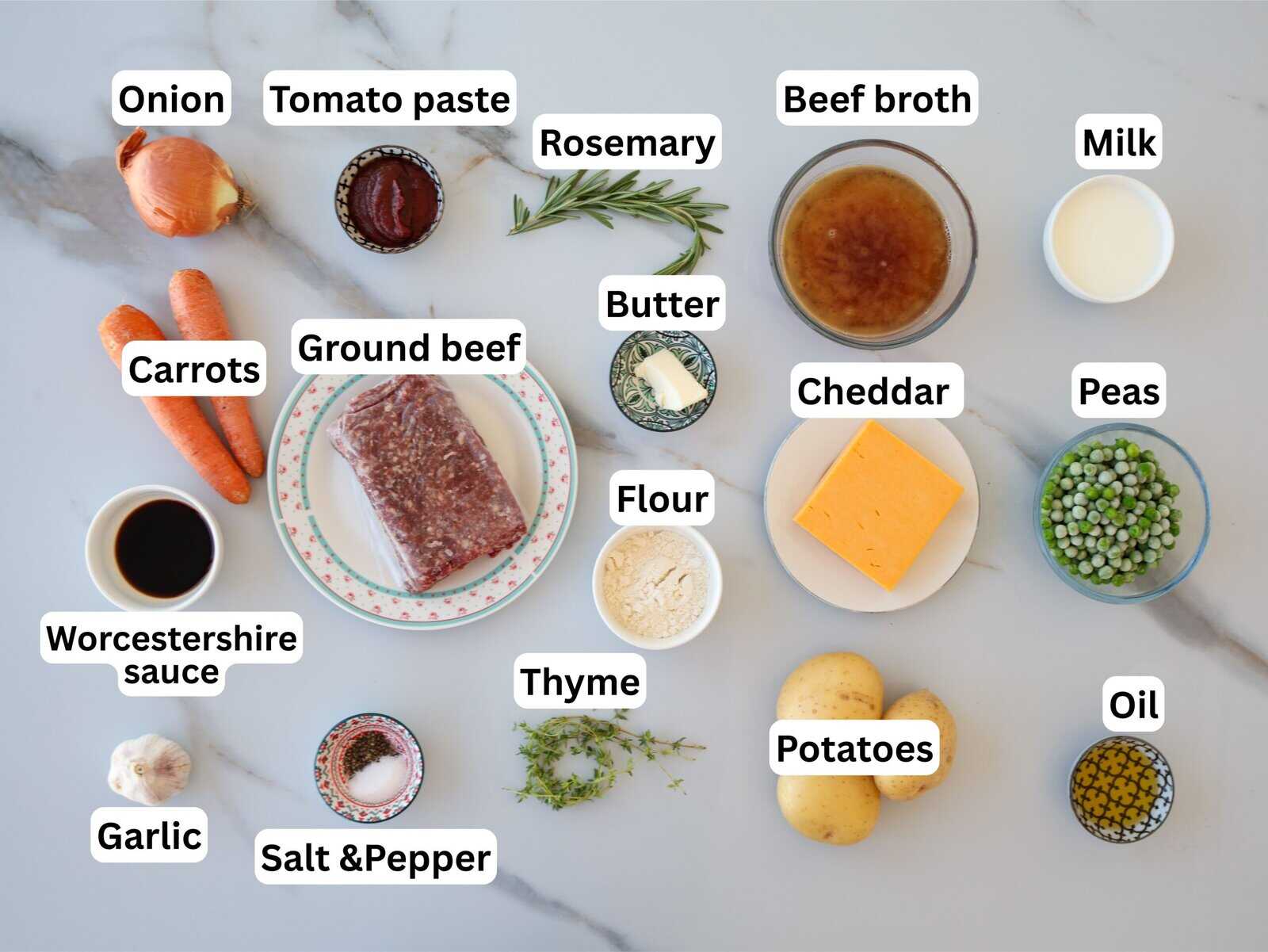 A variety of cottage pie ingredients are arranged on a white surface, each labeled: onion, tomato paste, rosemary, beef broth, milk, butter, cheddar, peas, oil, potatoes, thyme, salt & pepper, garlic, Worcestershire sauce, carrots, ground beef, and flour.