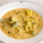 A close-up of a bowl of creamy broccoli and cheese soup, with a spoon lifting a portion. The soup contains visible broccoli pieces in a thick, cheesy broth.