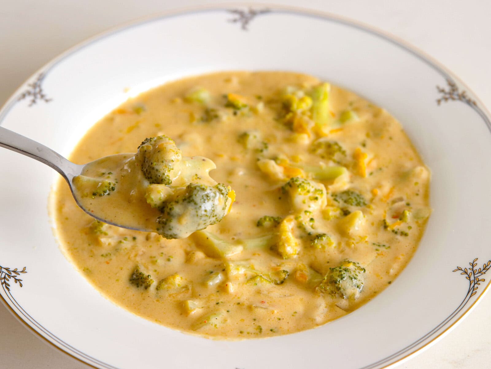 A close-up of a bowl of panera copycat broccoli and cheddar soup, with a spoon lifting a portion. The soup contains visible broccoli pieces in a thick, cheesy broth.