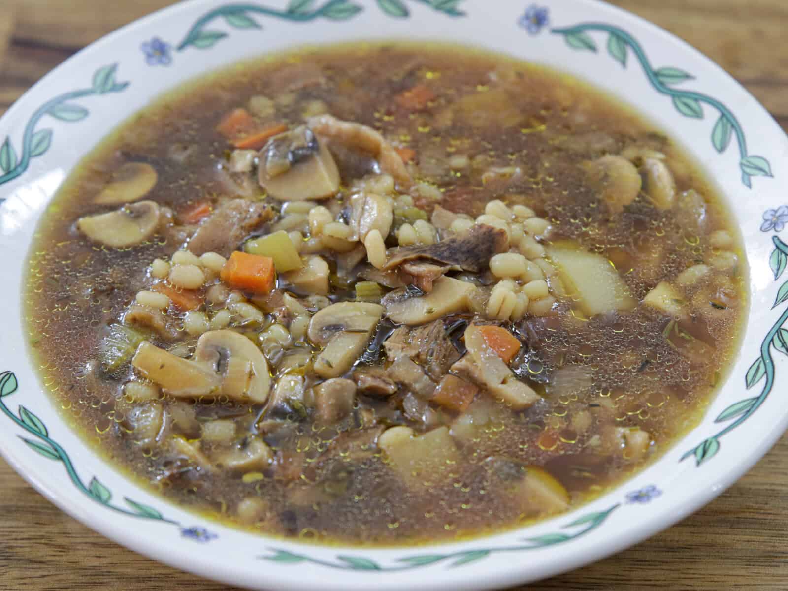 A bowl of mushroom barley soup with visible mushrooms, barley, carrots, potatoes, and broth, served in a white bowl with a green and blue floral pattern around the rim.