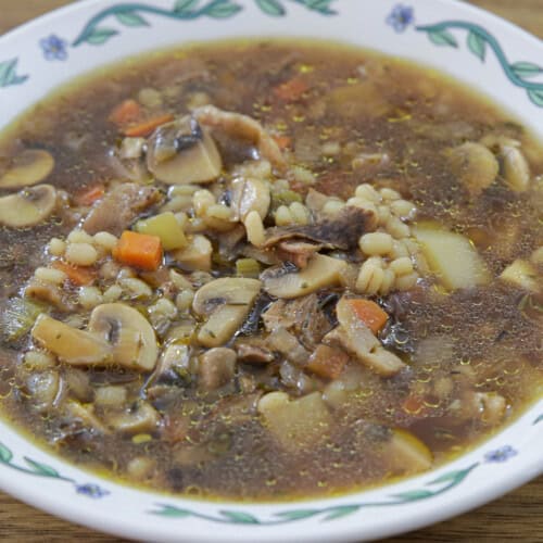 A bowl of mushroom barley soup with visible slices of mushrooms, diced carrots, potato chunks, barley, and herbs in a clear broth, served in a white bowl with a green and blue floral pattern.