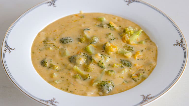 A white bowl with decorative trim filled with creamy broccoli cheddar soup, featuring chunks of broccoli and melted cheese in a thick, orange-tinted broth.