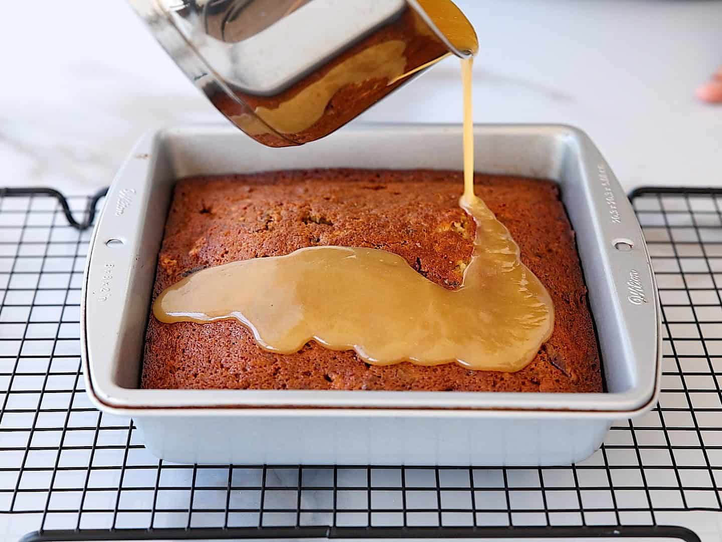 A golden sauce is being poured from a pot onto a freshly baked cake in a square metal pan, which is resting on a black wire cooling rack.