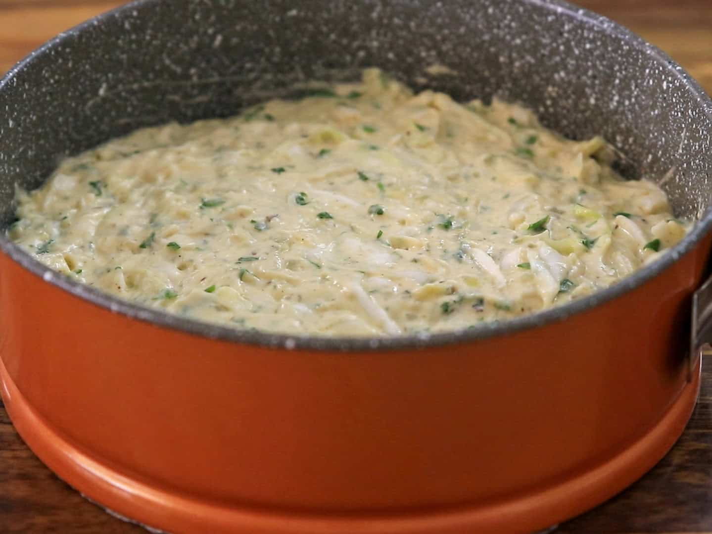 A round baking pan filled with uncooked batter, mixed with visible green herbs and shredded ingredients, sits on a wooden surface.