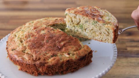A round, golden-brown savory cake with chopped herbs is on a white plate. A slice is being lifted, showing a moist, textured interior filled with green and white ingredients. The background is a wooden surface.