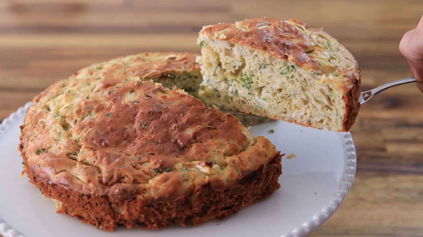 A round, golden-brown savory cabbage cake with chopped herbs is on a white plate. A slice is being lifted, showing a moist, textured interior filled with green and white ingredients. The background is a wooden surface.