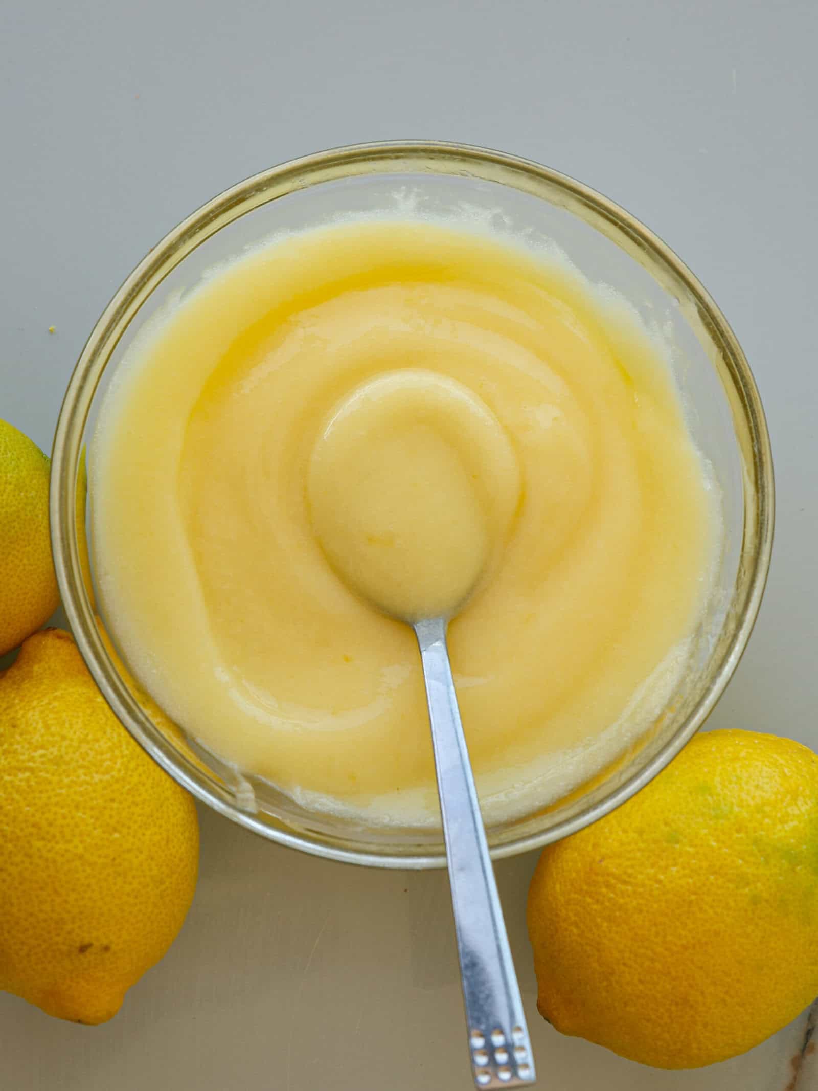 A glass bowl filled with creamy 4-ingredient lemon curd, with a spoon resting inside. Three whole lemons are placed around the bowl on a light surface.