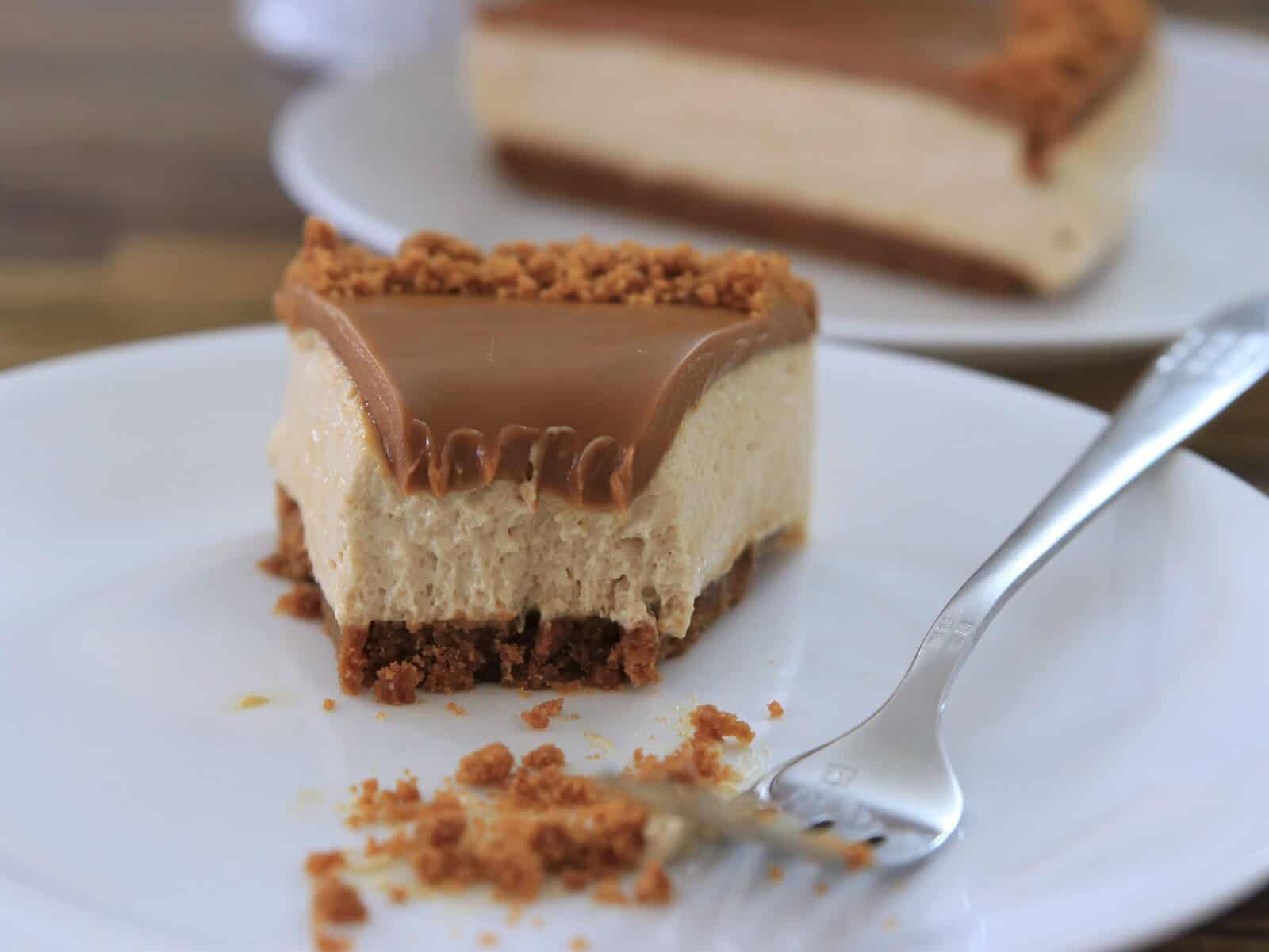A partially eaten slice of creamy biscoff cheesecake with a crumbly crust and a smooth caramel topping sits on a white plate beside a fork. Another full slice is visible in the background.