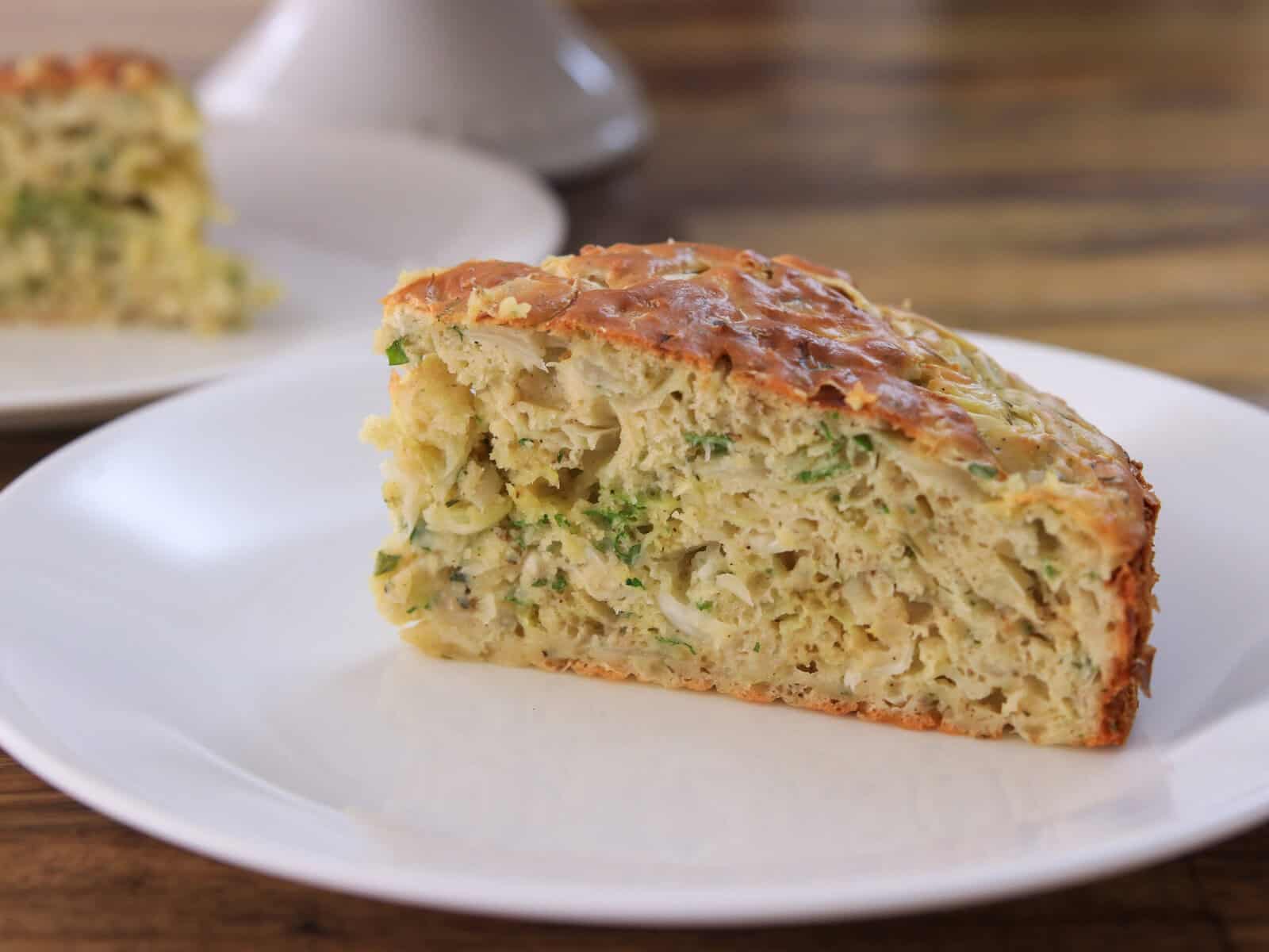 A slice of savory vegetable cake, showing a golden crust and a moist interior with visible pieces of zucchini and herbs, served on a white plate with a wooden table background.