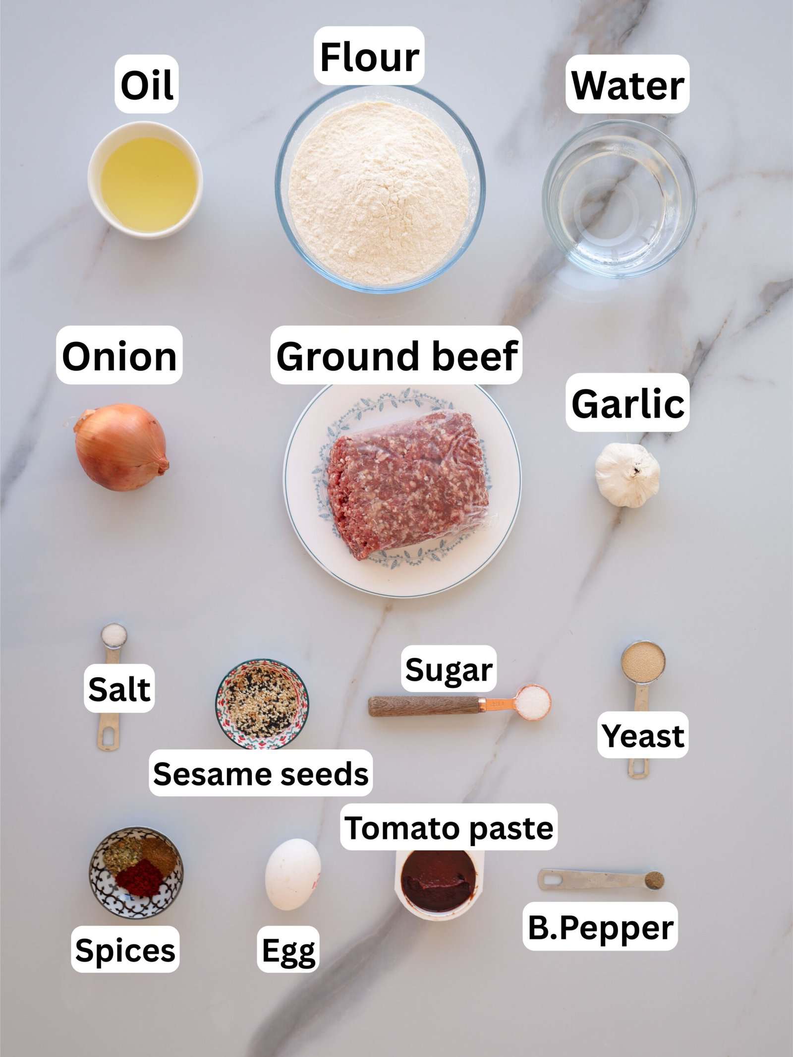 A marble surface displays labeled ingredients for a recipe: oil, flour, water, onion, ground beef, garlic, salt, spices, sesame seeds, tomato paste, sugar, egg, yeast, and black pepper.