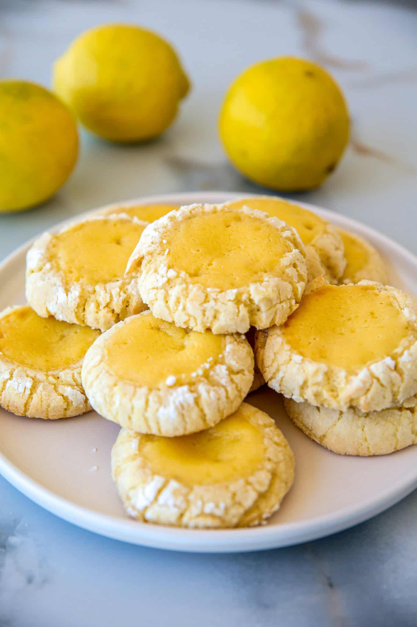 A white plate with a stack of crinkle-edged lemon curd cookies sits on a marble surface, with three whole lemons in the background.