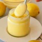 A spoonful of creamy lemon curd being lifted from a glass jar on a white plate, with fresh lemons in the background.